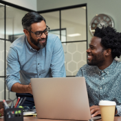 Two people in an office setting smiling and talking, one sitting at a desk with a laptop and coffee cup, the other standing and leaning over the desk.